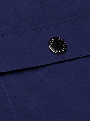 A close-up of the Shoda Cropped Wide-Leg Pant in dark blue nylon shows a flap with a round, black metal snap button engraved with text. Visible stitching and water-repellent fabric highlight the pant's quality craftsmanship.