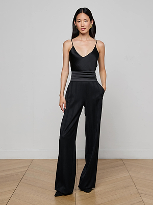 A woman with long, straight dark hair stands against a light gray wall, wearing L'AGENCE Minerva Cummerbund Trousers and a black satin sleeveless camisole, hands in pockets, gazing at the camera.