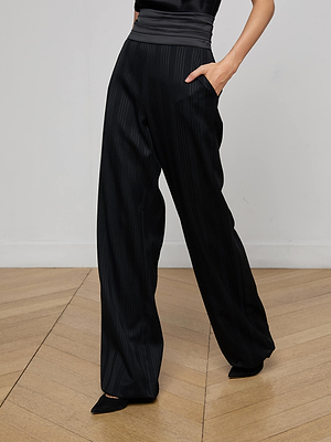 A person wearing L'AGENCE Minerva Cummerbund Trouser, featuring high-waisted, wide-leg shadow stripes and one hand in a pocket, stands on a light wooden floor against a plain white wall.