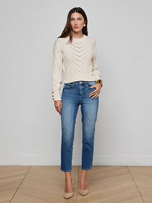 A woman with long brown hair wears the Celestia Slim-Leg Jean, a cream knit sweater, and beige heels, standing indoors against a light gray wall with neutral makeup and a confident posture.