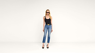 A woman with long blonde hair, wearing sunglasses, a black tank top, blue jeans, a belt, and black heels, stands against a plain white background.