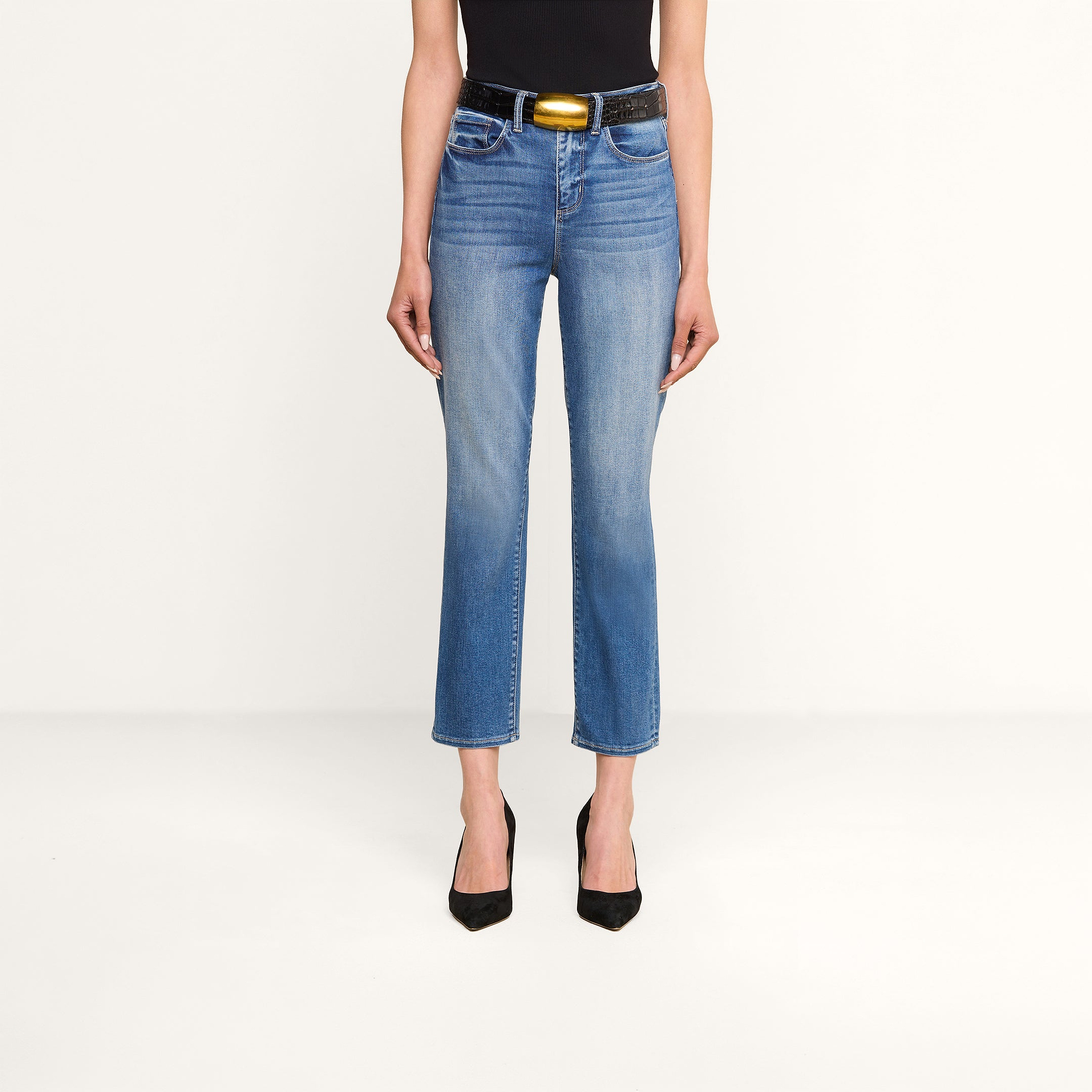 Woman wearing high rise Celestia Slim-Leg Jean in blue stretch denim, a black sleeveless top, a black belt with a gold buckle, and black pointed heels, standing against a plain white background.