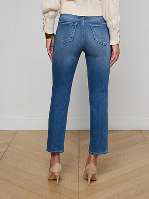 A person stands on a wooden floor facing away, wearing Celestia Slim-Leg Jean made of stretch denim, a cream sweater with buttoned cuffs, and beige high-heeled shoes. The jeans are blue, high rise, skinny fit with two back pockets.