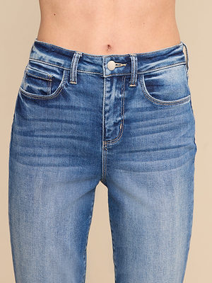 Un gros plan montre une personne portant le jean « Celestia Slim-Leg », un modèle à taille haute en denim stretch au délavage moyen, doté dune fermeture à bouton et à fermeture éclair. Ce jean présente de légers effets de délavage et de « whiskering », tandis que le ventre est visible au-dessus de la ceinture.