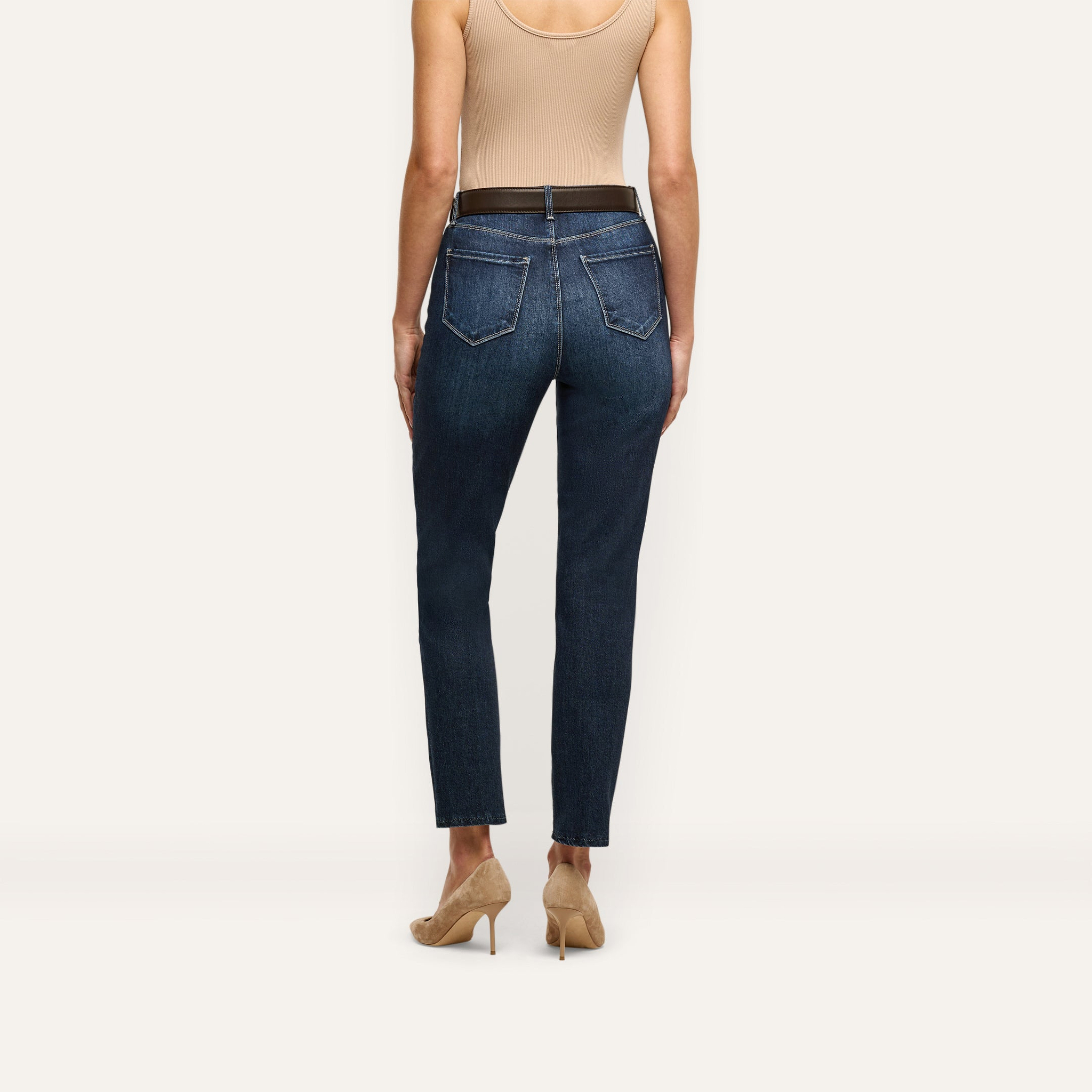 Back view of a person wearing Celestia Slim-Leg Jean ultra high rise, slim-leg stretch denim jeans, a tan tank top, a dark belt, and beige stiletto heels.