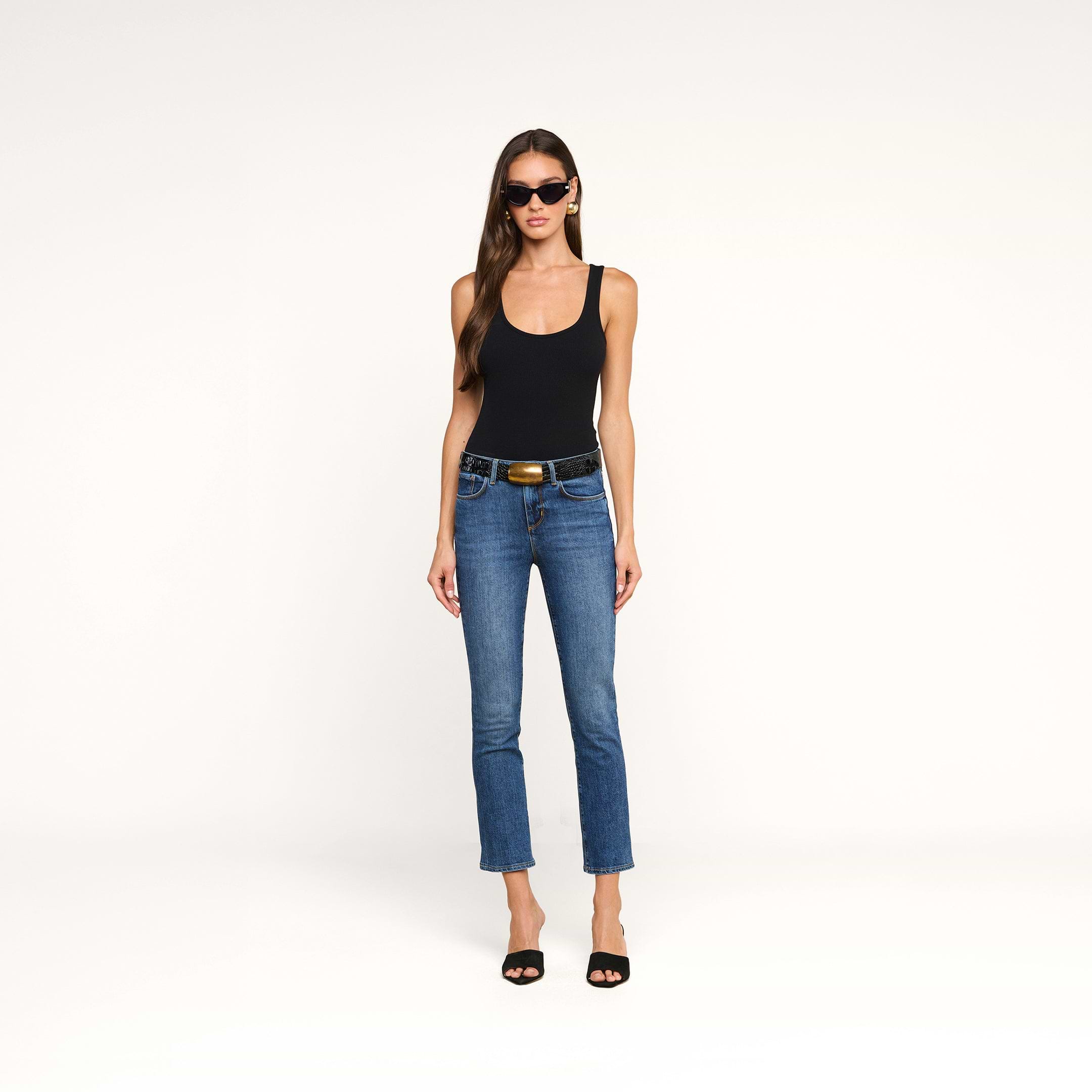 A woman stands against a plain white background wearing black sunglasses, a black tank top, the Celestia Slim-Leg Jean with a belt, and black heels.
