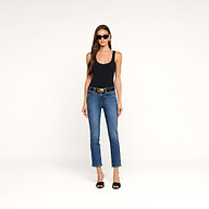 A woman stands against a plain beige background, facing forward with a neutral expression. She is wearing sunglasses, a black tank top, black heels, and the Celestia Slim-Leg Jean in an indigo wash paired with a belt.