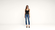 A woman wearing sunglasses, a black tank top, blue jeans, and black heels stands against a plain white background, facing the camera.