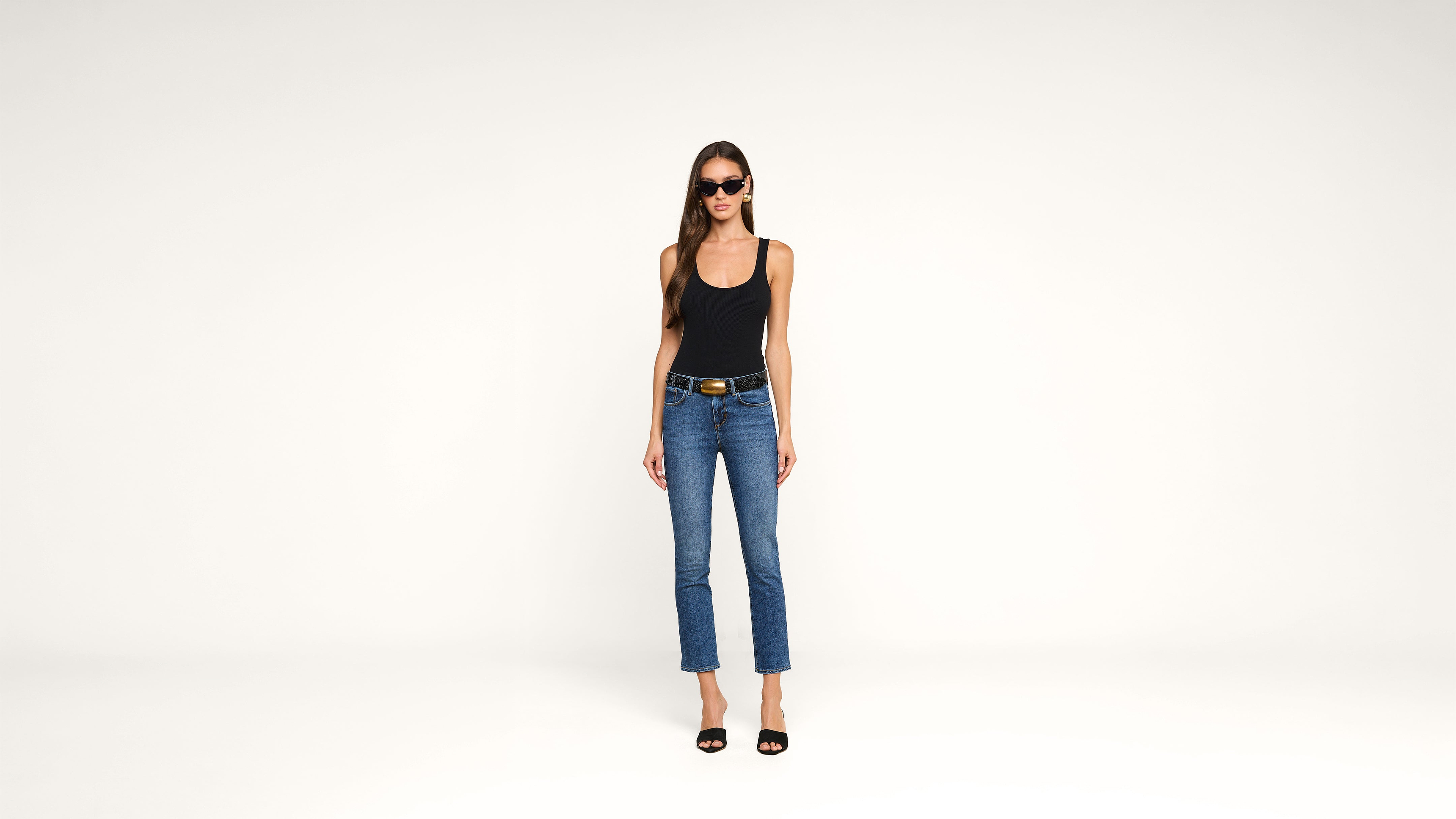 A woman wearing sunglasses, a black tank top, blue jeans, and black heels stands against a plain white background, facing the camera.