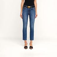 A person wearing the Celestia Slim-Leg Jean in a rich indigo wash, styled with a black belt featuring a gold buckle, a black sleeveless top, and black open-toe heels stands against a beige background.