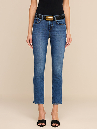 A person wearing the Celestia Slim-Leg Jean in a rich indigo wash, styled with a black belt featuring a gold buckle, a black sleeveless top, and black open-toe heels stands against a beige background.
