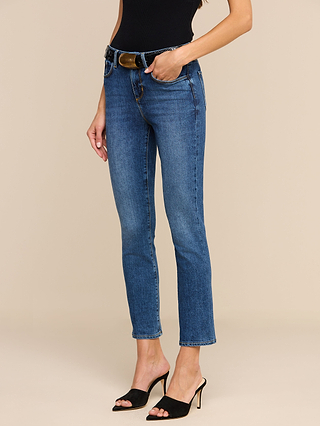 Wearing the Celestia Slim-Leg Jean in an indigo wash, crafted from stretch denim, a woman pairs the high-rise skinny jeans with a black sleeveless top and black heeled sandals, standing with one hand in her pocket against a beige background.