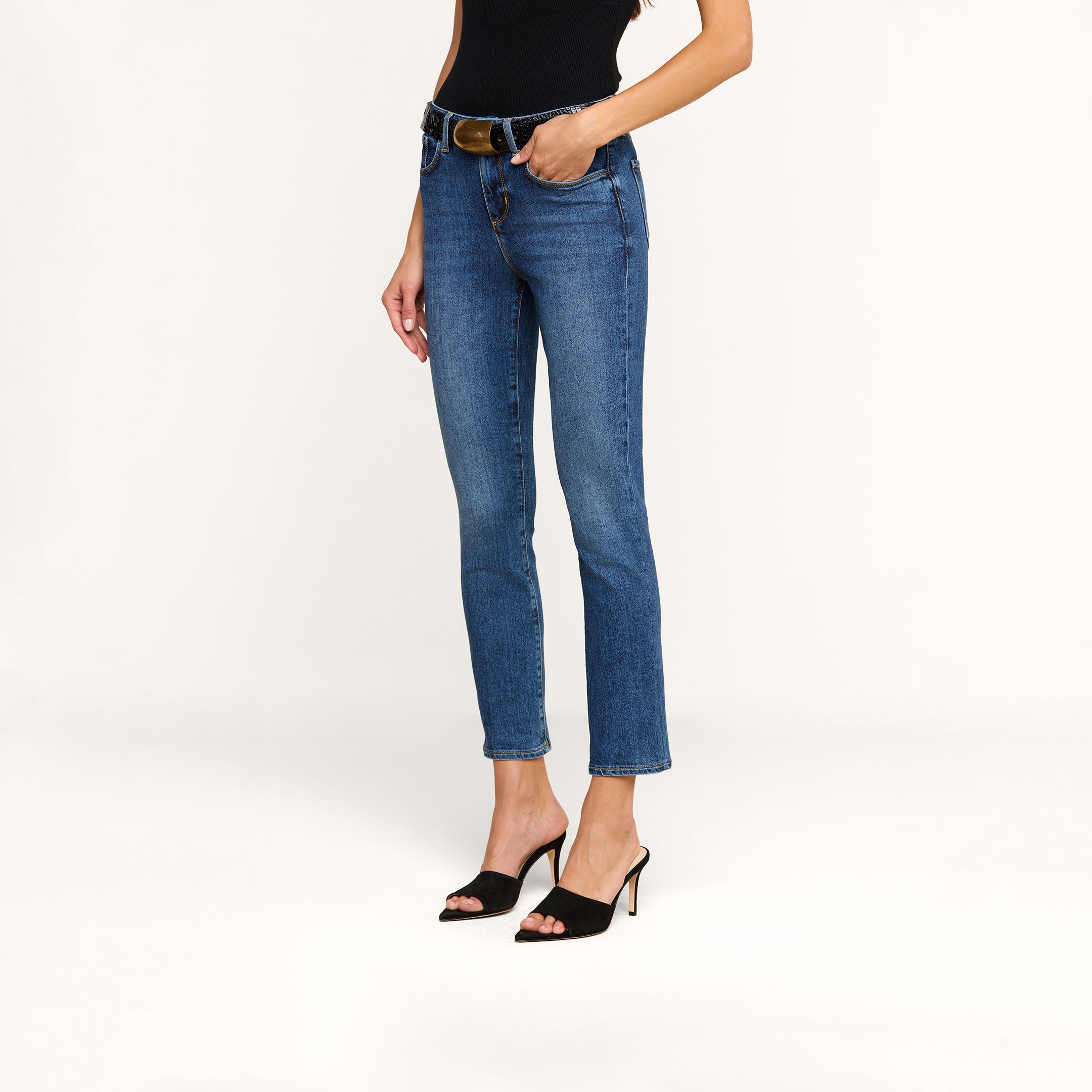 Wearing the Celestia Slim-Leg Jean in an indigo wash, crafted from stretch denim, a woman pairs the high-rise skinny jeans with a black sleeveless top and black heeled sandals, standing with one hand in her pocket against a beige background.