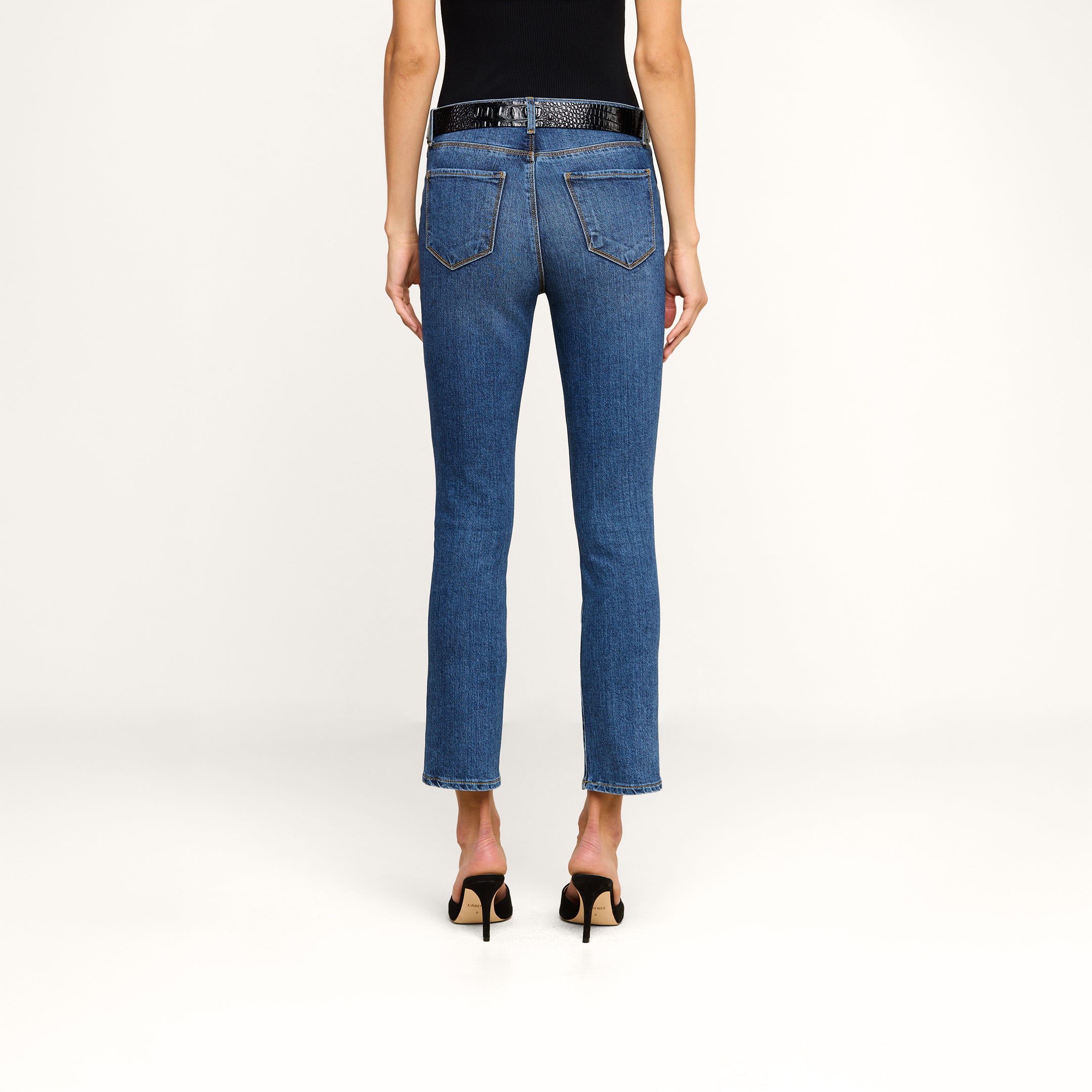 A person is shown from the back against a plain beige background, wearing Celestia Slim-Leg Jeans in an indigo wash, a black belt, and black heeled sandals.