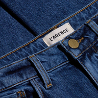 Close-up of blue, indigo wash Celestia Slim-Leg Jean showing a metal button, belt loops, and a white label with “L’AGENCE” printed in black text. The fabric texture and stitching are clearly visible on these high-rise jeans crafted from comfortable stretch denim.