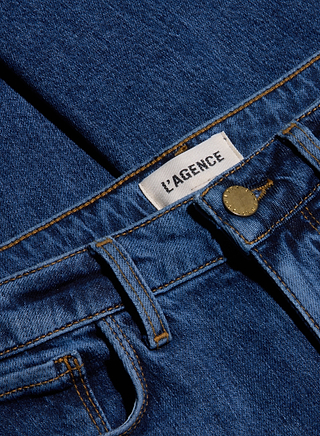 Close-up of blue, indigo wash Celestia Slim-Leg Jean showing a metal button, belt loops, and a white label with “L’AGENCE” printed in black text. The fabric texture and stitching are clearly visible on these high-rise jeans crafted from comfortable stretch denim.