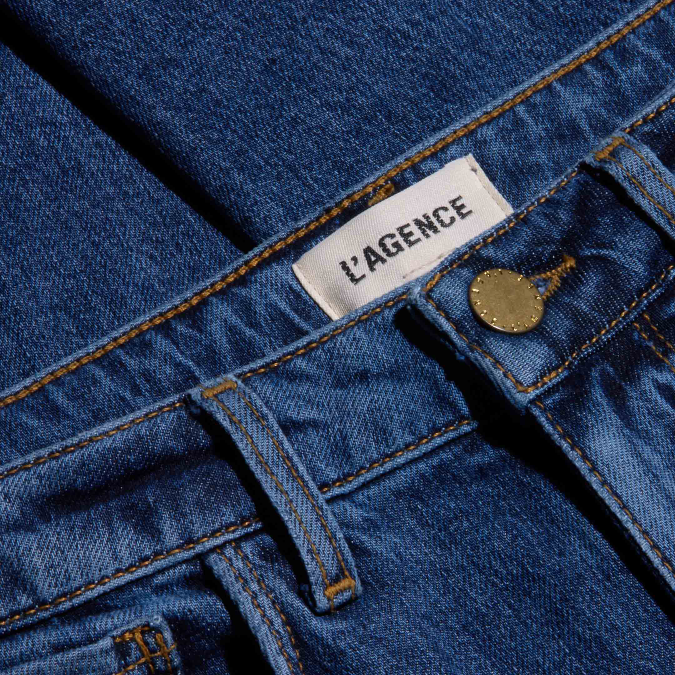 Close-up of blue, indigo wash Celestia Slim-Leg Jean showing a metal button, belt loops, and a white label with “L’AGENCE” printed in black text. The fabric texture and stitching are clearly visible on these high-rise jeans crafted from comfortable stretch denim.