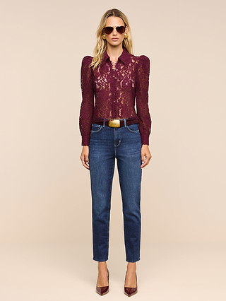 A woman stands against a beige background wearing sunglasses, a burgundy lace long-sleeve blouse, the Celestia Slim-Leg Jean in a dark indigo wash with a brown belt, and burgundy heels.