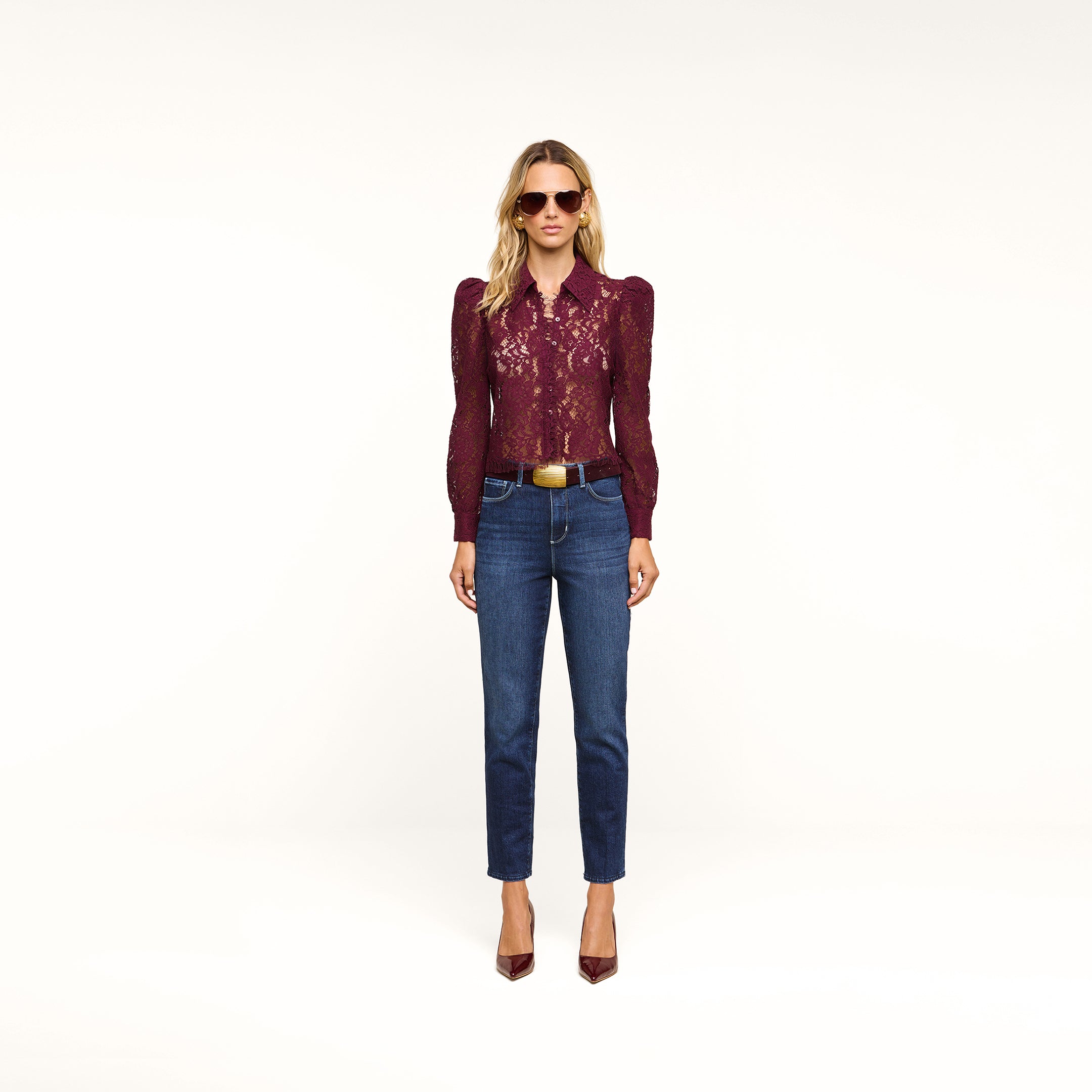 A woman stands against a beige background wearing sunglasses, a burgundy lace long-sleeve blouse, the Celestia Slim-Leg Jean in a dark indigo wash with a brown belt, and burgundy heels.