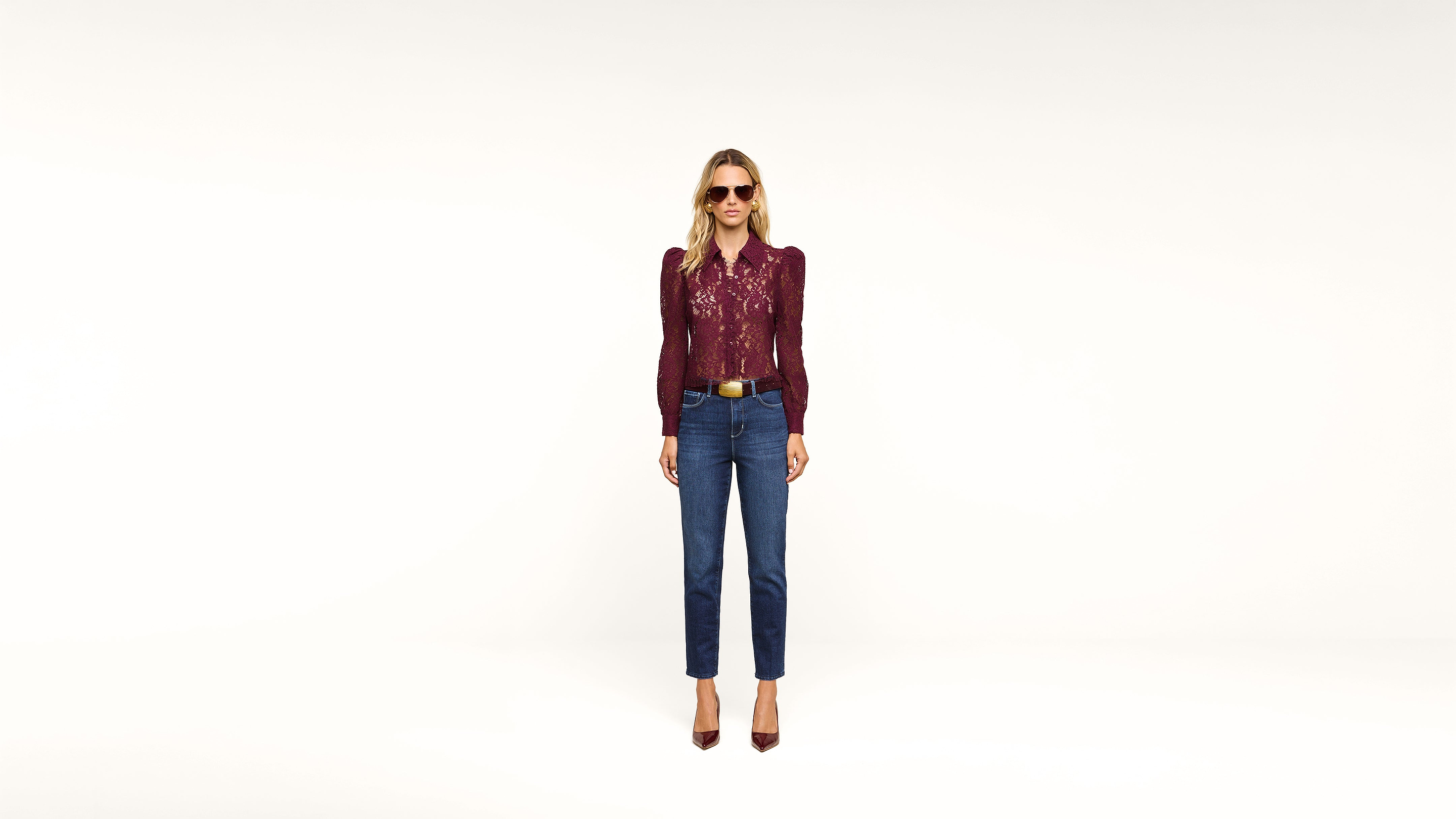 A woman wearing sunglasses, a burgundy long-sleeve top, blue jeans, and burgundy heels stands against a plain white background.