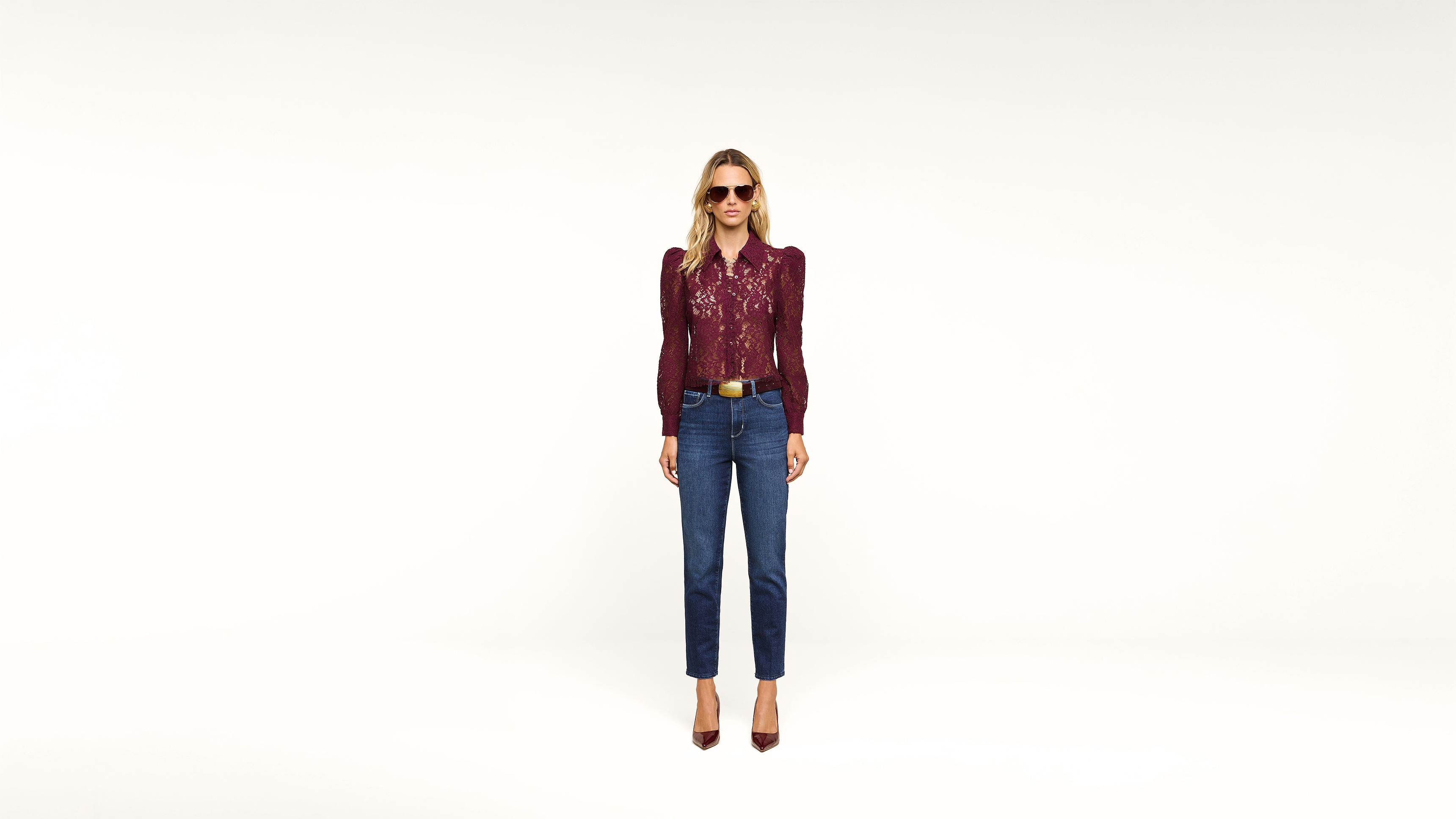 A woman wearing sunglasses, a burgundy long-sleeve top, blue jeans, and burgundy heels stands against a plain white background.