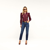 A woman stands against a plain beige background wearing sunglasses, a burgundy lace top with puffed sleeves, the Celestia Slim-Leg Jean in an ultra high rise dark indigo wash and classic stretch denim, paired with a tan belt and burgundy heels.