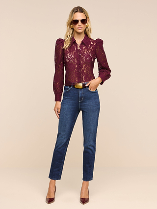 A woman stands against a plain beige background wearing sunglasses, a burgundy lace top with puffed sleeves, the Celestia Slim-Leg Jean in an ultra high rise dark indigo wash and classic stretch denim, paired with a tan belt and burgundy heels.