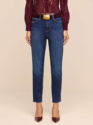 A person stands against a beige background, wearing the Celestia Slim-Leg Jean in a classic dark indigo stretch denim, paired with a burgundy lace top and matching burgundy heels; only the lower half of their body is visible.