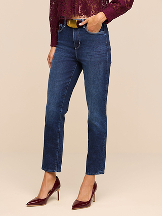 Wearing the Celestia Slim-Leg Jean in classic stretch denim, paired with a maroon lace top and maroon high heels, the person stands with one hand in their pocket against a plain beige background.