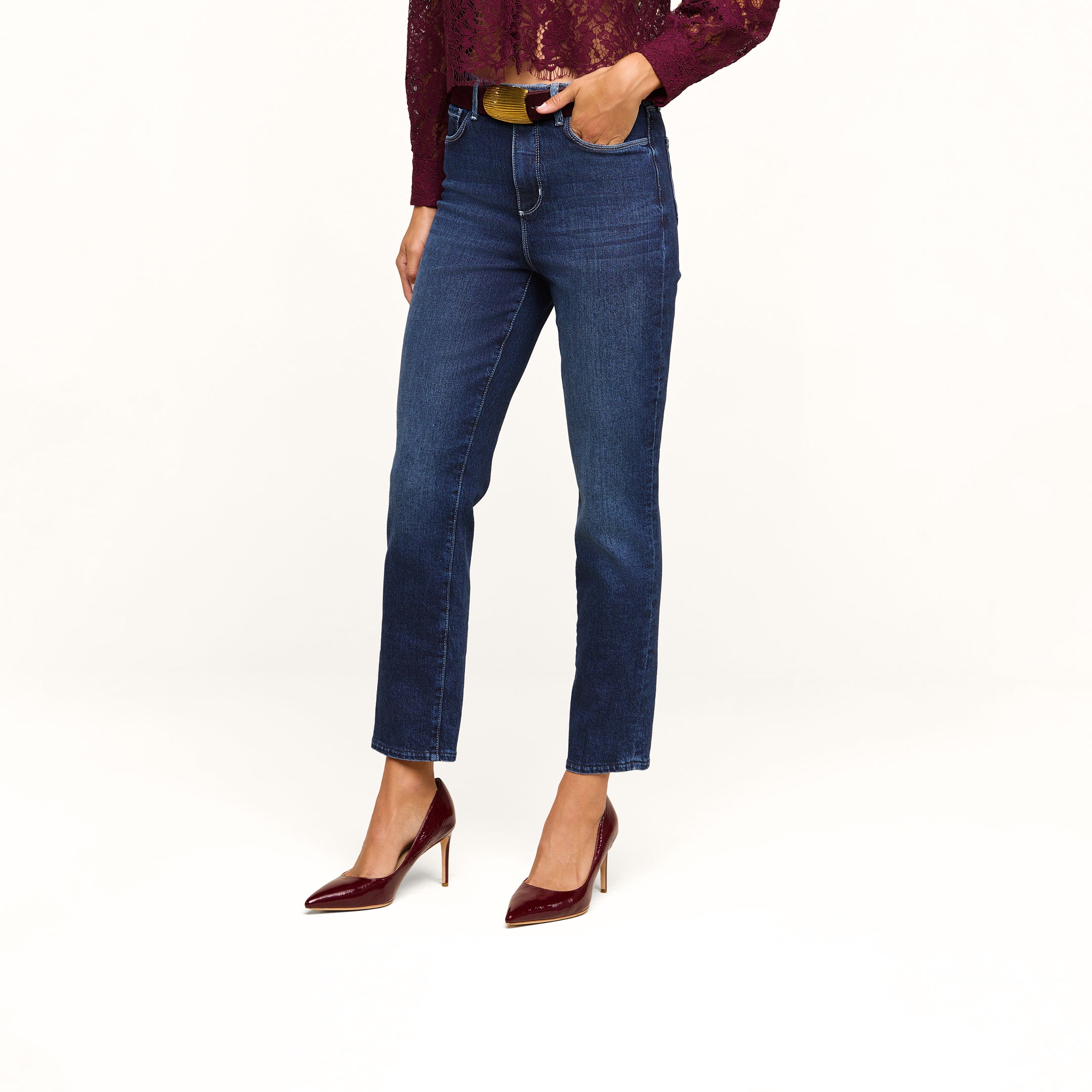 Wearing the Celestia Slim-Leg Jean in classic stretch denim, paired with a maroon lace top and maroon high heels, the person stands with one hand in their pocket against a plain beige background.