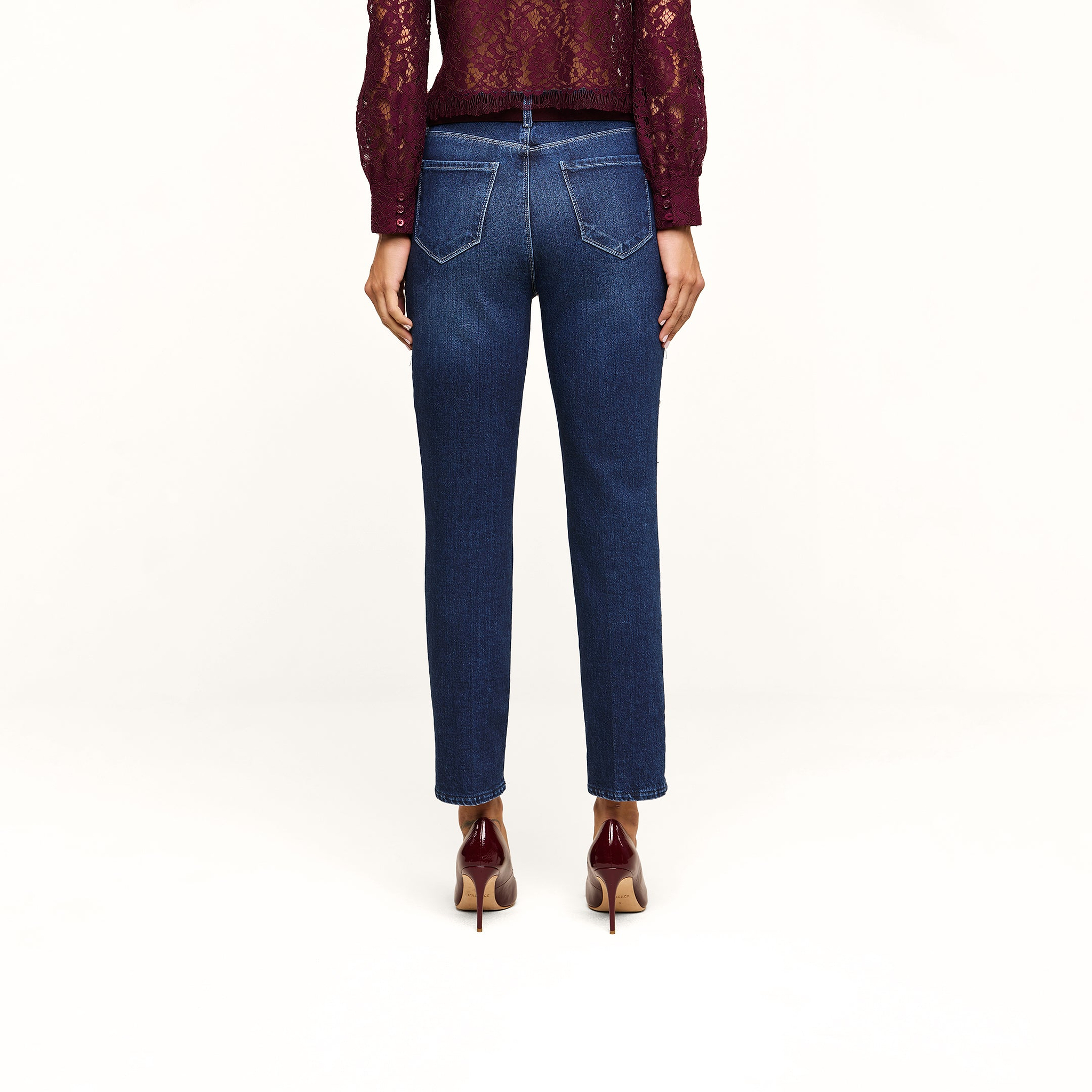 A person wearing the Celestia Slim-Leg Jean in a dark indigo wash, paired with a burgundy lace top and matching burgundy heels, stands facing away to display the back view of their outfit against a plain beige background. The classic stretch denim of the Celestia Slim-Leg Jean offers both comfort and style.