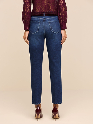 A person wearing the Celestia Slim-Leg Jean in a dark indigo wash, paired with a burgundy lace top and matching burgundy heels, stands facing away to display the back view of their outfit against a plain beige background. The classic stretch denim of the Celestia Slim-Leg Jean offers both comfort and style.