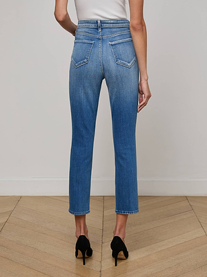 Someone models the L'AGENCE Celestia Slim-Leg Jean, featuring a high rise and two-tone blue design, paired with black high heels. They stand on a light wood floor facing away, set against a plain, light background.