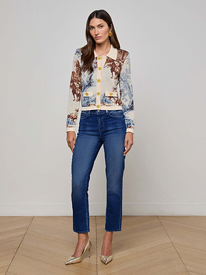 A woman with long brown hair stands indoors on a wooden floor, wearing L'AGENCE Celestia Slim-Leg Jean in vintage wash blue, a patterned button-up cardigan with brown and blue designs, and silver heels, looking at the camera.