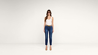 A woman with long hair wears sunglasses, a white tank top, blue jeans with a belt, and tan heels, standing against a plain white background.