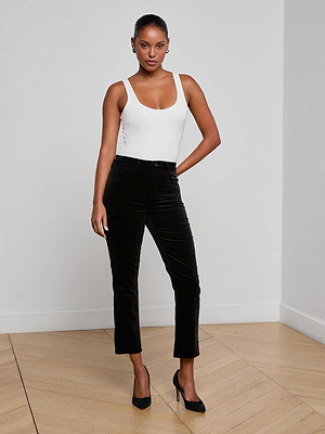 A woman stands in a light room with wooden floors, wearing the L'AGENCE Celestia Velvet Slim-Leg Jean, a white sleeveless top, and black heels, her hand on her hip.