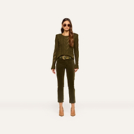 A woman wears the Celestia Velvet Slim-Leg Jean in dark green, paired with an olive green sweater, a brown belt, and beige heels. She has long wavy hair, sunglasses, and stands against a plain light background.