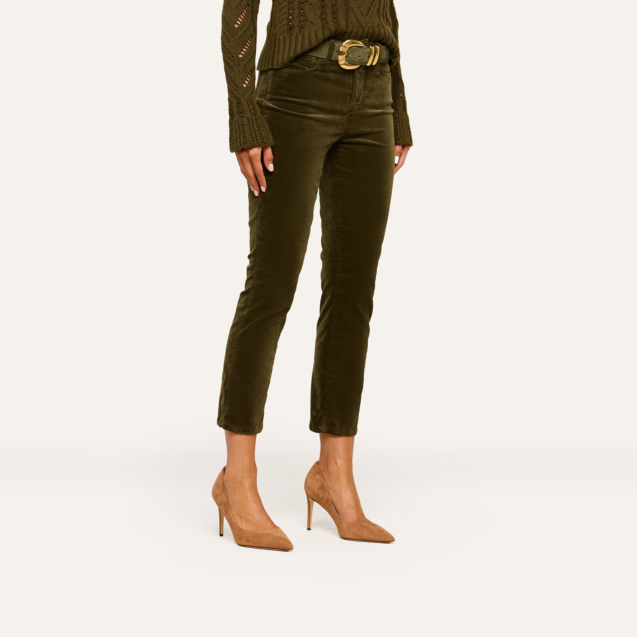 A person wearing an olive green sweater, the Celestia Velvet Slim-Leg Jean with a belt, and tan high heels stands against a plain background.