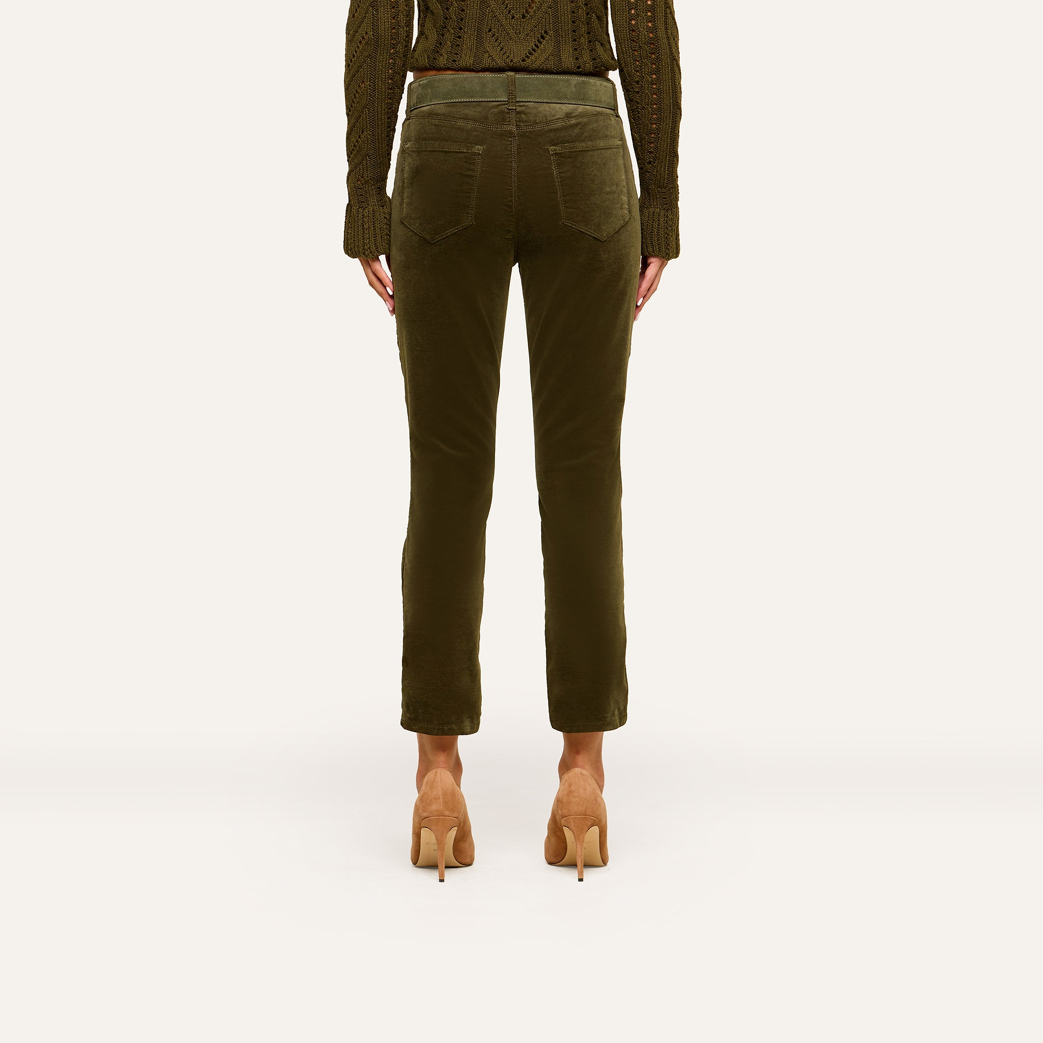 A person stands facing away against a plain background, showcasing the back view of the outfit featuring the Celestia Velvet Slim-Leg Jean in dark green, paired with a matching knit sweater and tan high heels.
