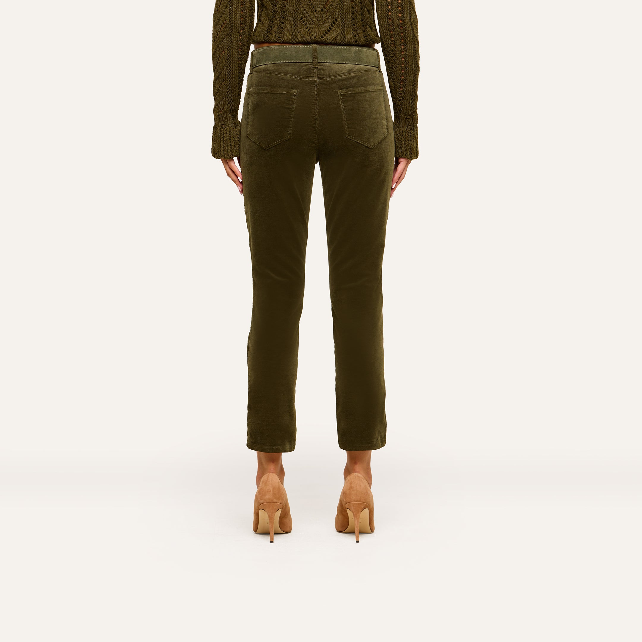 A person stands facing away against a plain background, showcasing the back view of the outfit featuring the Celestia Velvet Slim-Leg Jean in dark green, paired with a matching knit sweater and tan high heels.