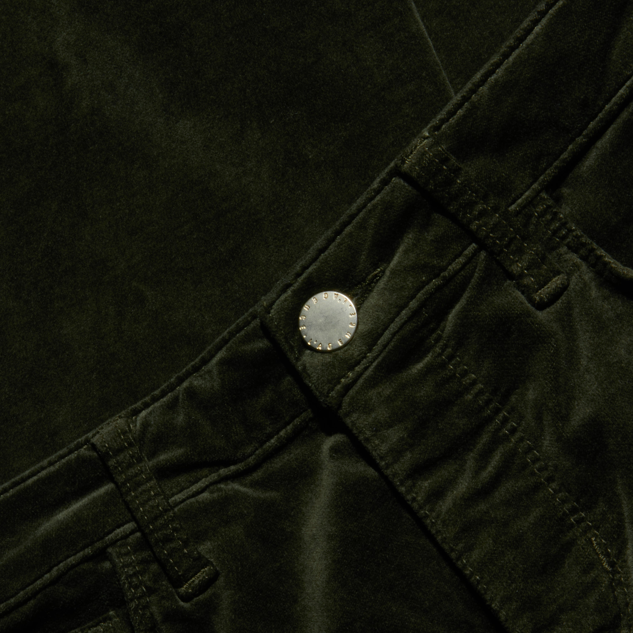 Close-up of the Celestia Velvet Slim-Leg Jean in dark green, featuring the waistband, belt loops, and metallic button, with emphasis on the texture and details of the stretch velvet fabric and stitching.