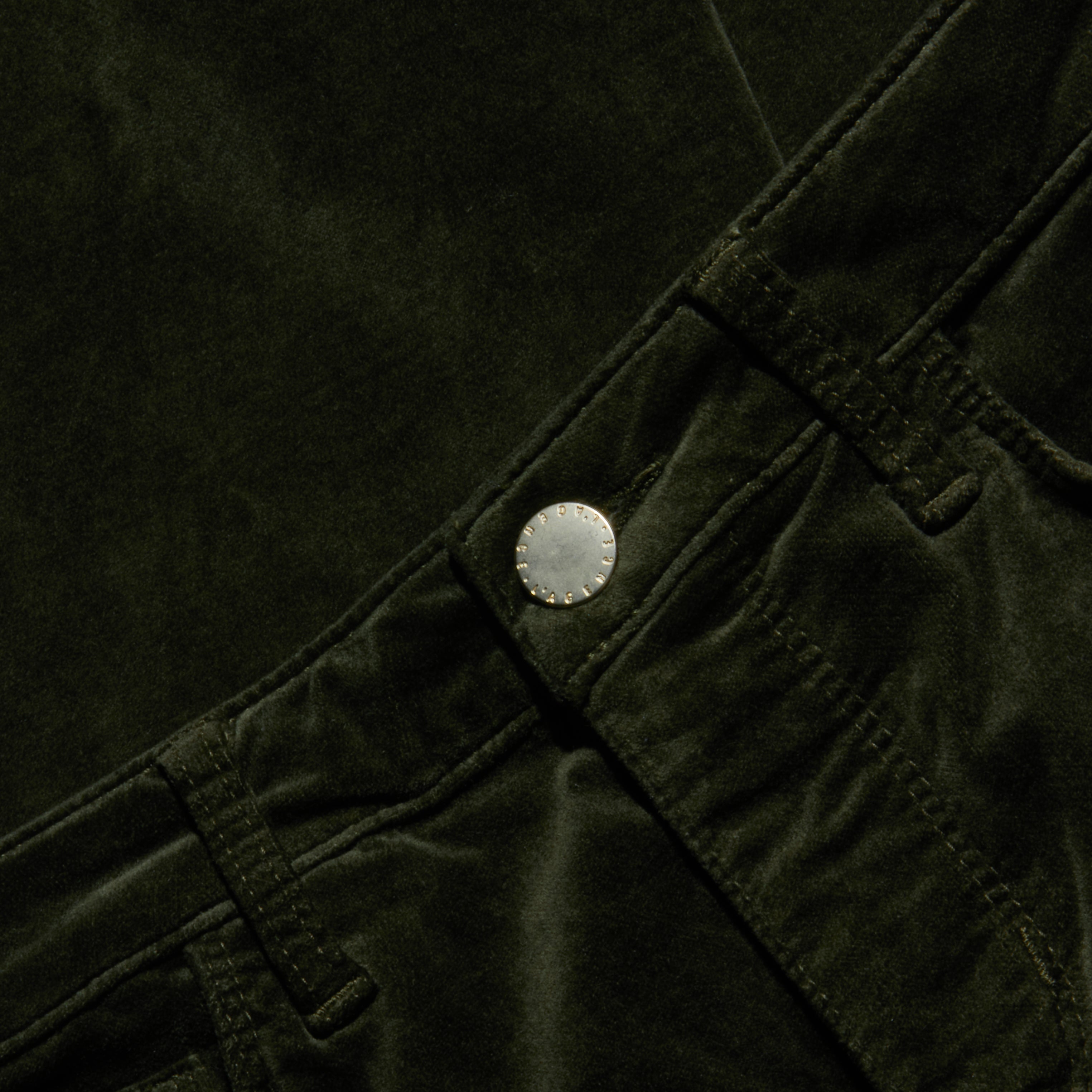 Close-up of the Celestia Velvet Slim-Leg Jean in dark green, featuring the waistband, belt loops, and metallic button, with emphasis on the texture and details of the stretch velvet fabric and stitching.