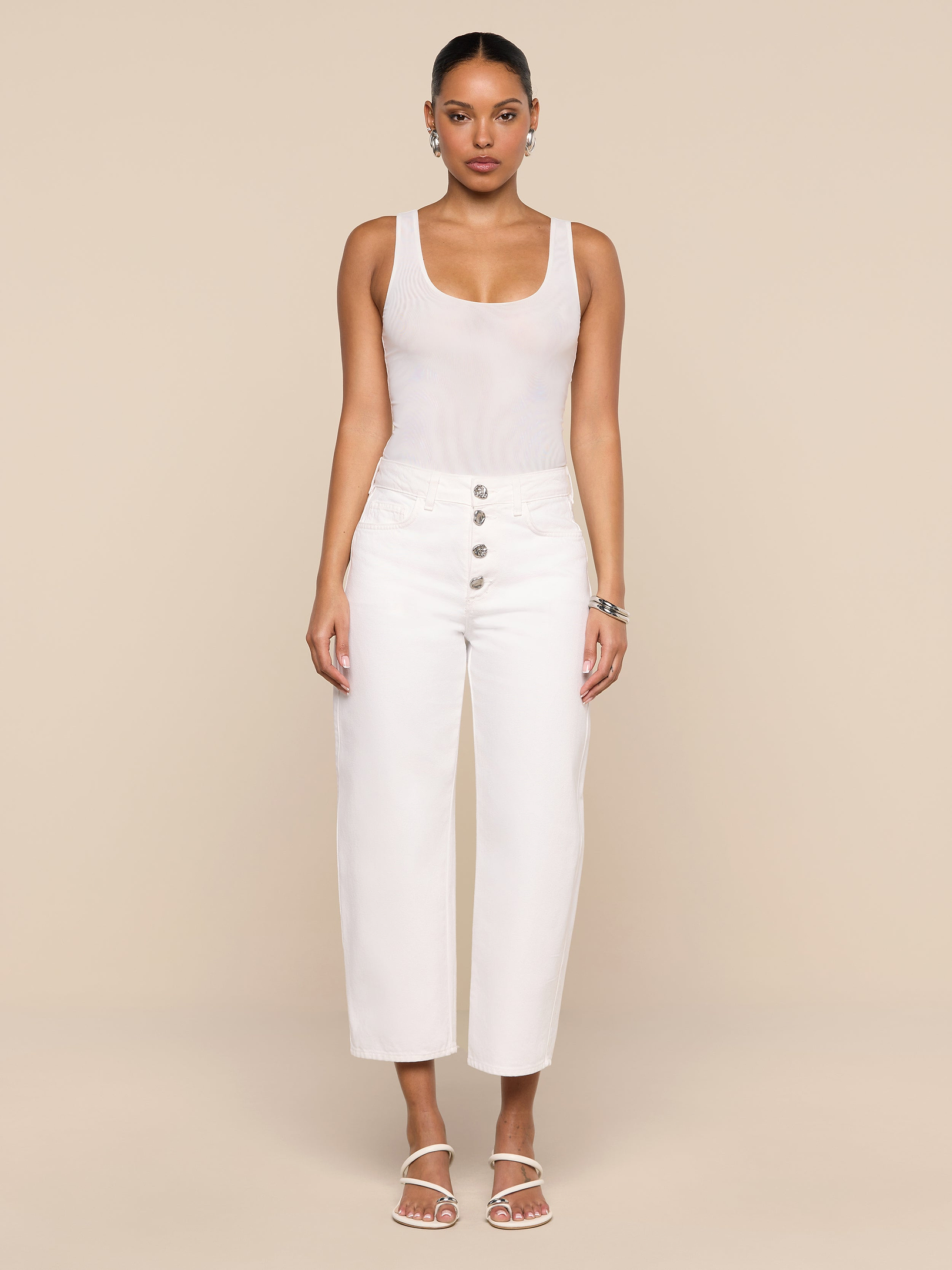 A woman stands facing forward against a plain white background, wearing a sleeveless white top, the Liora Cropped Jean—high-rise, white wide-leg pants with buttons—white sandals, and hoop earrings.