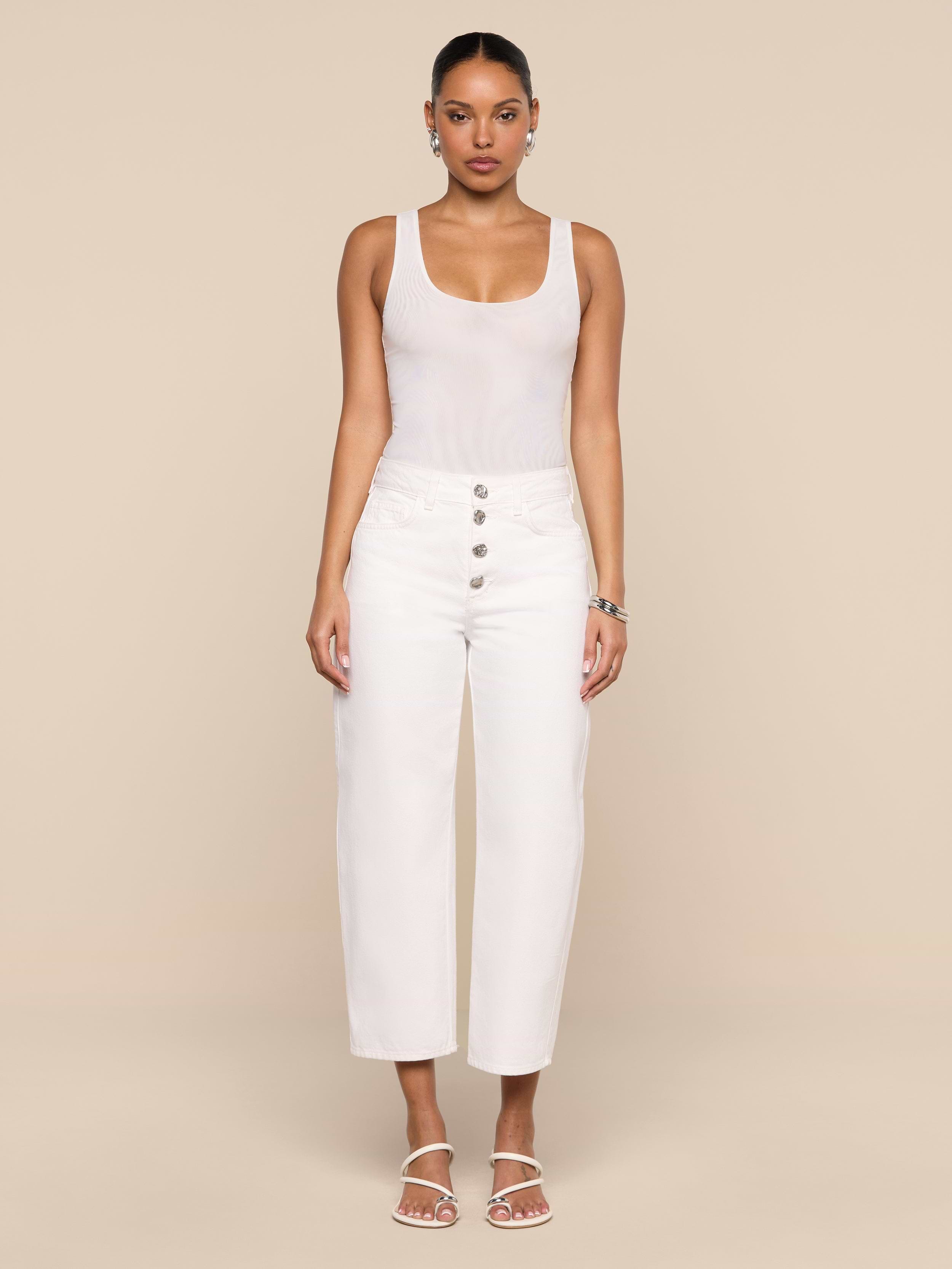 A woman stands facing forward against a plain white background, wearing a sleeveless white top, the Liora Cropped Jean—high-rise, white wide-leg pants with buttons—white sandals, and hoop earrings.