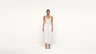 A woman stands facing forward against a plain white background, wearing a sleeveless white top, high-waisted white pants with buttons, and white sandals.