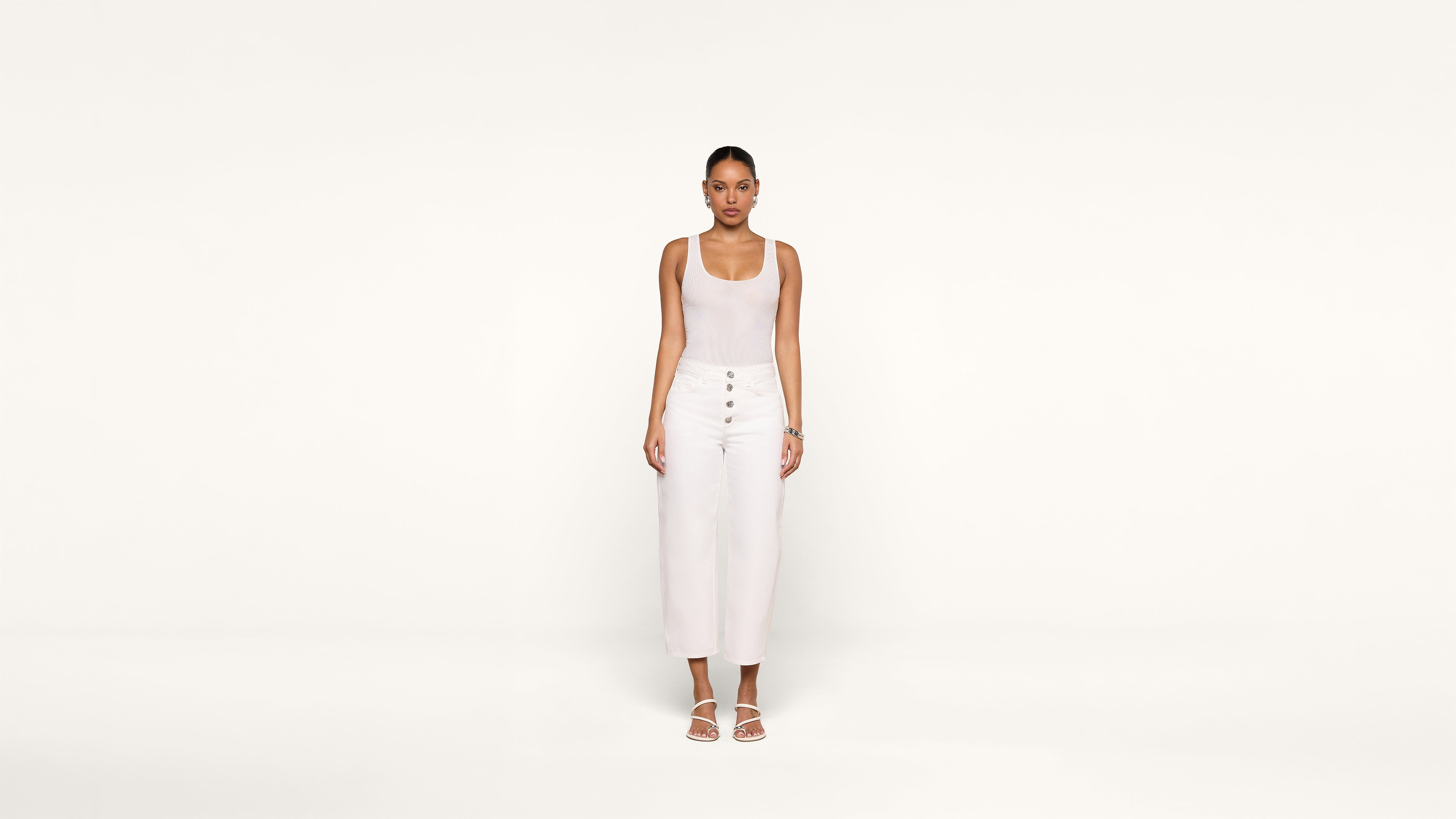 A woman stands facing forward against a plain white background, wearing a sleeveless white top, high-waisted white pants with buttons, and white sandals.