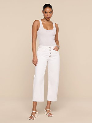 A woman stands against a beige background, wearing a sleeveless white tank top, the Liora Cropped Jean—high rise and wide leg with buttons—white heeled sandals, and silver hoop earrings. Her hair is tied back and her hand is in her pocket.
