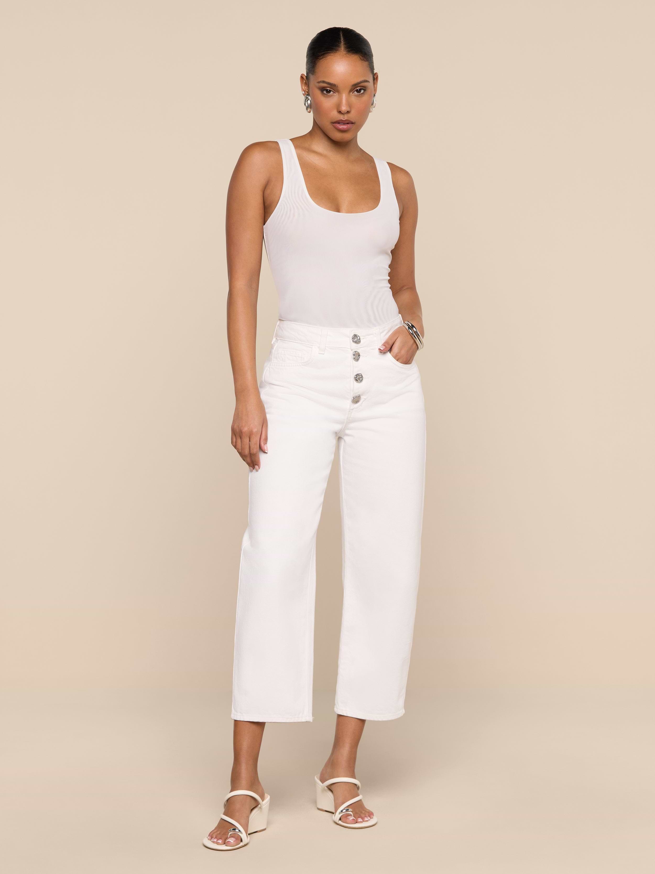A woman stands against a plain white background wearing a sleeveless beige top, the Liora Cropped Jean—high-rise, wide-leg white pants with button details—and white sandals. Her hands are in her pockets and she has a neutral expression.