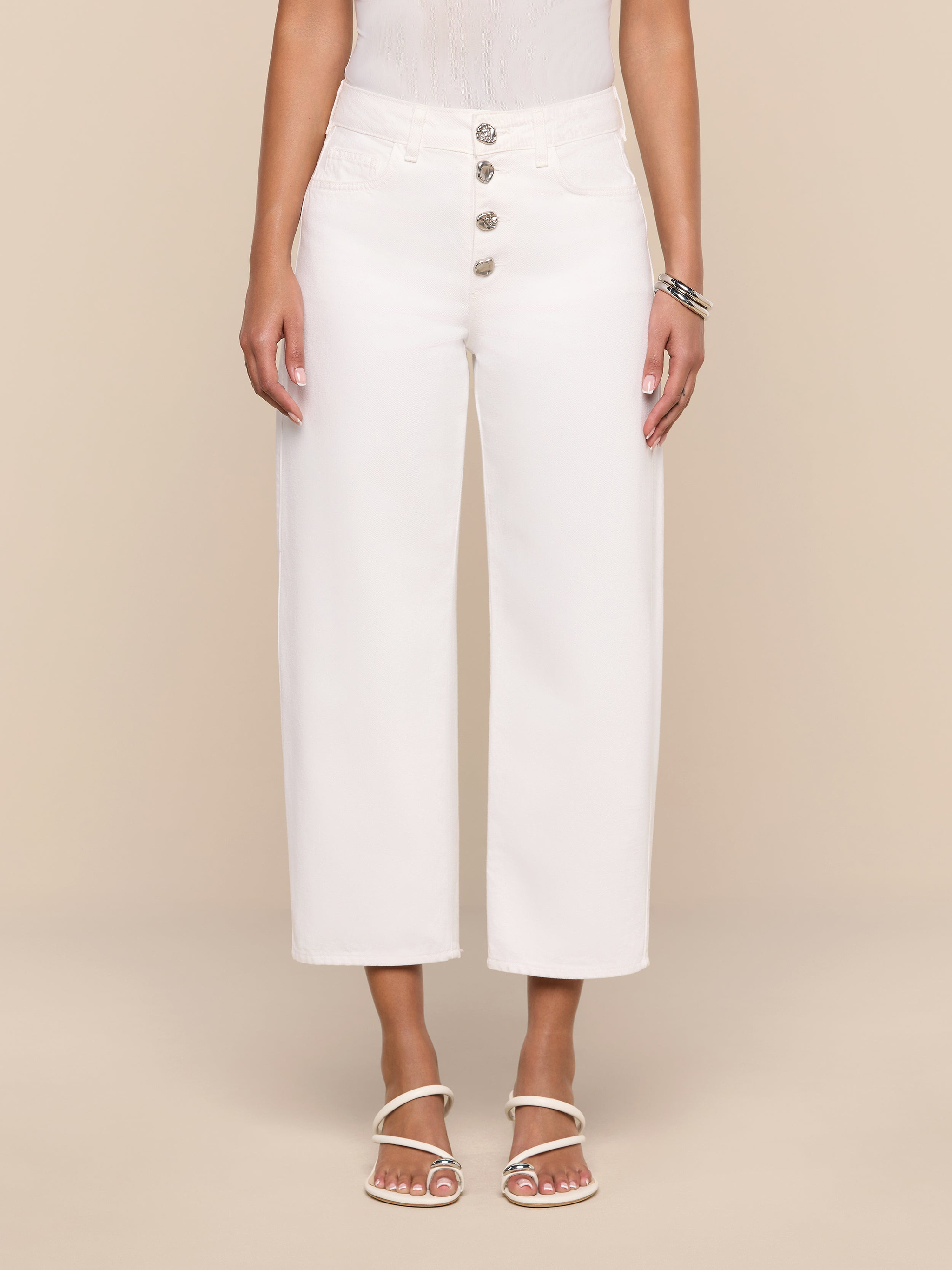 A woman wearing the Liora Cropped Jean—white high-rise, wide-leg jeans with large buttons—paired with a white sleeveless top, white heeled sandals, and a bracelet, stands against a plain white background. Her face is out of frame.