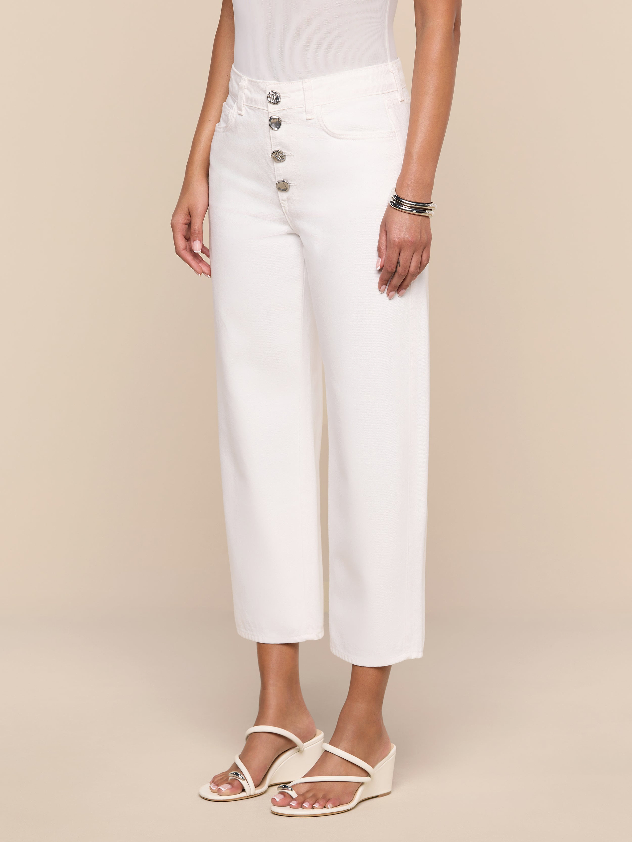 Wearing the Liora Cropped Jean—a pair of high-rise, wide-leg white jeans with large silver buttons—a person stands against a plain white background, styled with a sleeveless white top, white sandals, and silver bracelets.